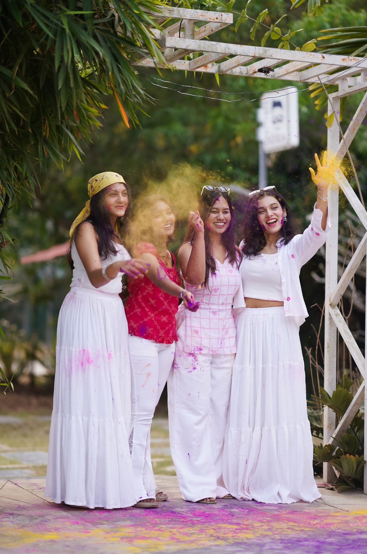 Four women in white and colorful outfits standing outdoors with a decorative arch and greenery.