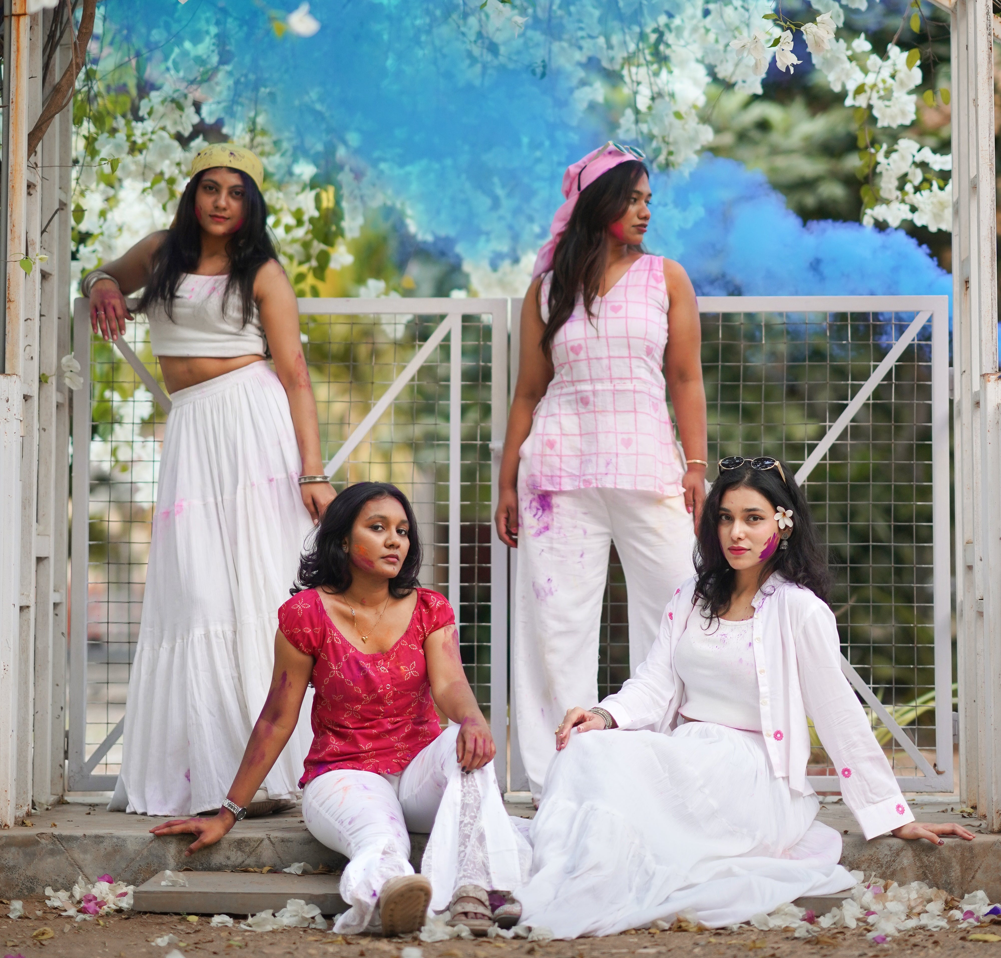 Four women in traditional attire standing under a floral archway with a blue sky in the background.