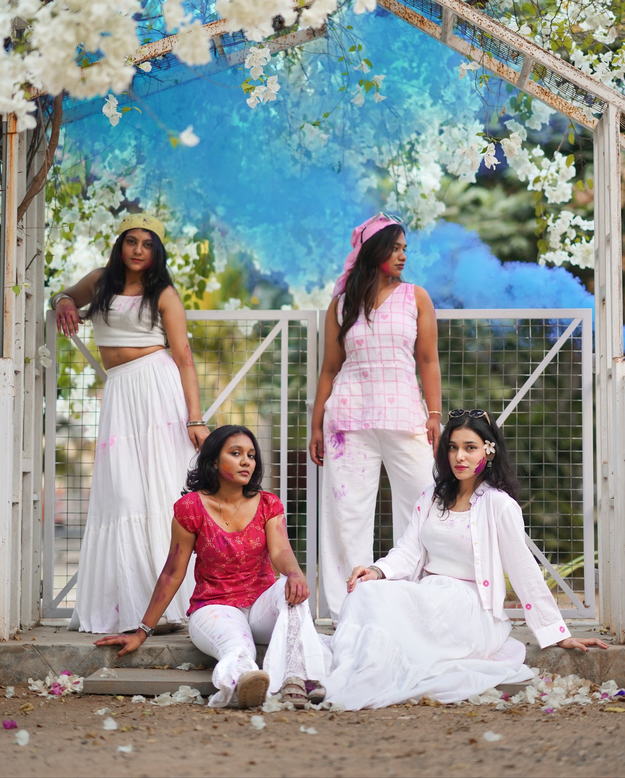 Four women in traditional attire standing under a floral archway with a blue sky in the background.