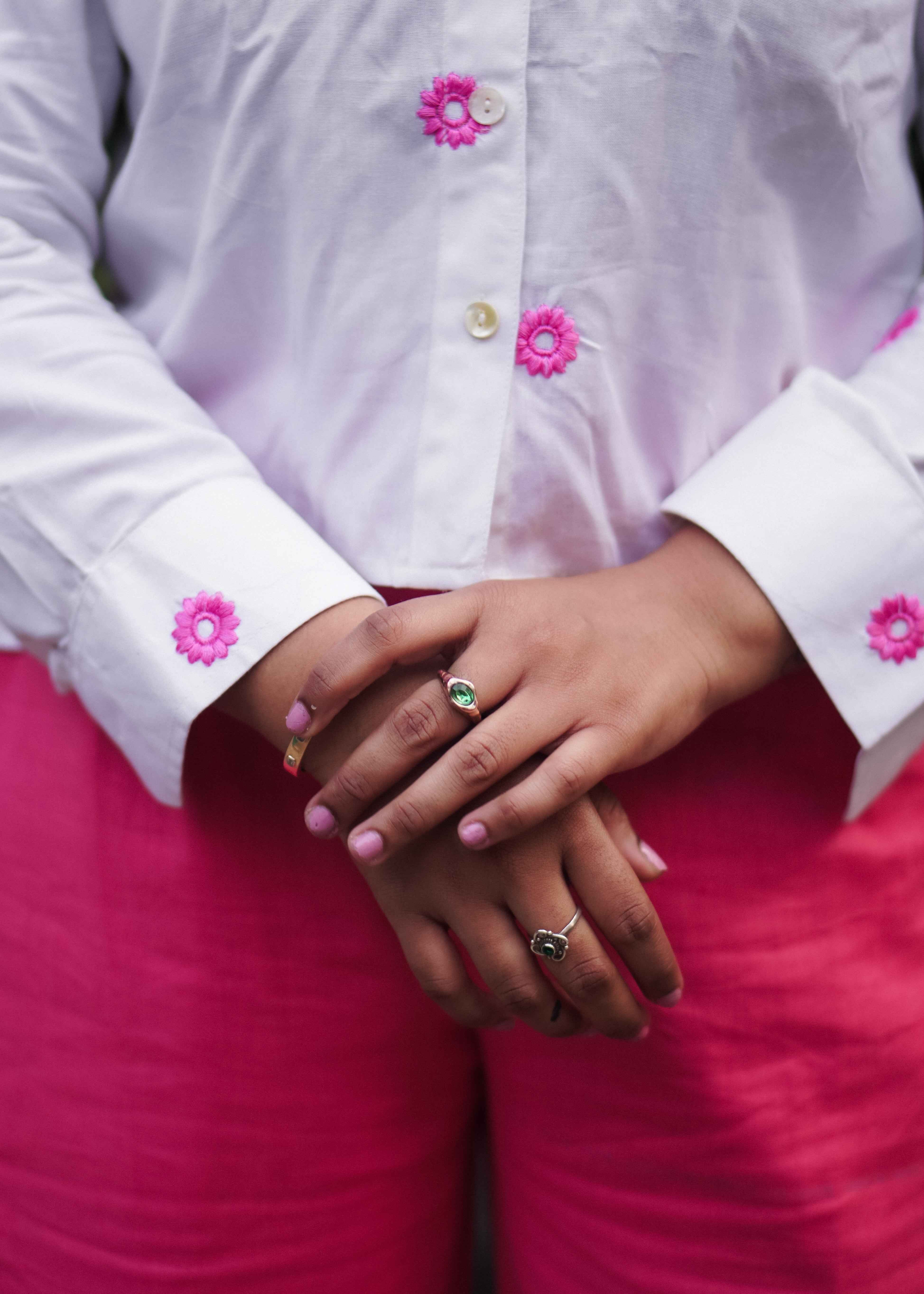 Close-up of hands with pink nail polish wearing rings, against a white shirt with floral buttons.