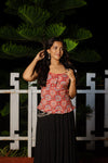 Woman wearing a red patterned top and black skirt standing in front of a white fence with green foliage. pop naari